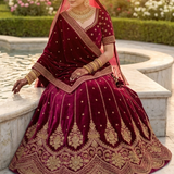 Bride seated in rosewood maroon bridal lehenga with gold embroidery, side profile by a fountain