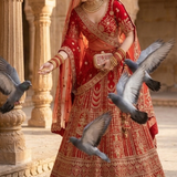 Bride in crimson bridal lehenga standing amid pigeons in a heritage courtyard.