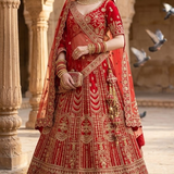 Bride in crimson red bridal lehenga with gold embroidery, standing in a heritage courtyard