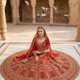 Bride seated with crimson bridal lehenga skirt spread in a full circle, showing gold embroidery.