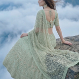 Woman seated in sage green embroidered lehenga on a cliff edge above clouds