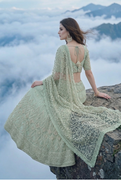 Woman seated in sage green embroidered lehenga on a cliff edge above clouds