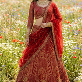 Bride in scarlet red bridal lehenga with gold embroidery, standing in a wildflower field